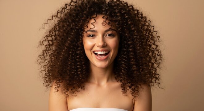 Happy woman with voluminous curly hair smiling at the camera. Beauty portrait with a focus on natural hairstyle and hair care concept. Isolated on a beige studio background with copy space