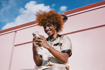 Smiling young Black man with curly red afro sitting outdoors, holding smartphone in both hands....