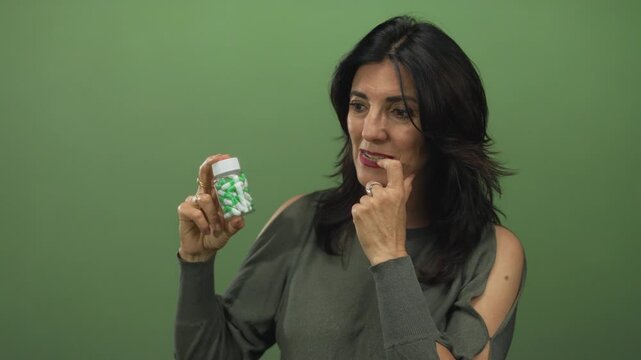 Middle aged hispanic woman holds green pill bottle and hand on chin in studio; concern health uncertainty.