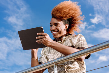 Young African American man with curly ginger afro smiling at digital tablet, holding stylus, wearing patterned shirt, representing technology-driven creativity, innovation, and freelance culture.