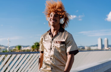 Fototapeta premium Stylish man with curly ginger afro and wireless headphones walking confidently, touching his hair, symbolizing individuality, personal technology, and the joy of music in motion.