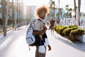 Young man with curly afro carrying backpack, boxing gloves, and wireless headphones outdoors. Lifestyle photo symbolizing active routine, mobility, and digital lifestyle.