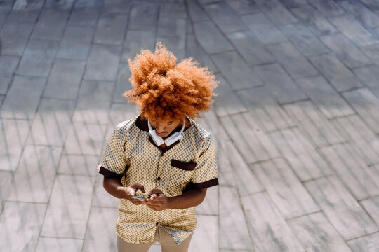 Young man with curly afro, wearing headphones on neck, using smartphone outdoors. Represents digital lifestyle, portable connectivity, and the role of mobile devices in daily expression.