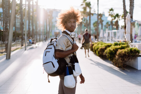 Young man with curly afro carrying backpack, boxing gloves, and wireless headphones outdoors. Lifestyle photo symbolizing active routine, mobility, and digital lifestyle. - Powered by Adobe