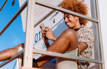 Smiling young man in patterned shirt using smartphone outdoors, leaning back casually. Conceptual stock photo of mobile connectivity, lifestyle, and self-expression through technology.