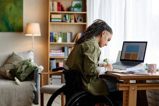 Young adult woman with disability sitting in wheelchair working at desk using laptop and writing in notebook in home office setting, focused on completing remote work tasks