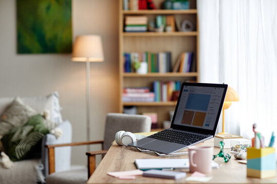 Modern workspace featuring open laptop on wooden desk with wireless headphones, notebooks, pen, and sticky notes, bookshelf and armchair visible in softly lit background