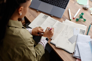 Young adult woman working at desk reviewing documents with QR codes, holding pen and wearing smartwatch, laptop and office supplies scattered on wooden surface