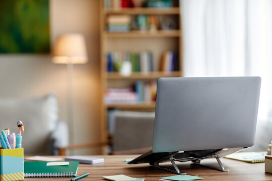 Modern laptop standing on desk in home office with blurred bookshelf and lamp in background, workspace organized with stationery and notebooks, no people visible