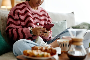 Young adult Caucasian woman sitting on sofa using smartphone, wearing smartwatch and necklace, holding device with both hands, food and coffee visible in foreground, casual home setting