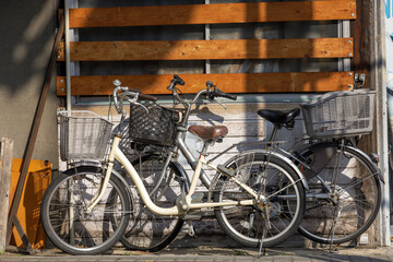 Two vintage-style Japanese Mama-chari bikes with baskets parked together on narrow sidewalk under dappled sunlight next to wooden panels covering window of small everyday urban home.