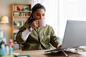 Young adult woman sitting at desk using laptop and drinking from mug, wearing smartwatch, working...