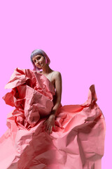A model showcases artistic flair, elegantly draped in crumpled pink paper against a bold pink backdrop. The playful scene highlights creativity and fashion, blending textures and hues