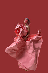 A talented model stands gracefully, embodying modern fashion in a stunning dress crafted from crumpled pink paper. The vivid red backdrop enhances the artistic vibe, highlighting creativity