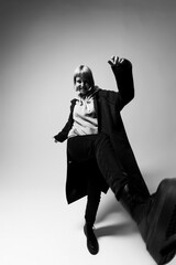 A person with light hair energetically kicks one leg forward, wearing a cozy hoodie and a long coat. The minimalist background highlights their joyful expression and dynamic movement