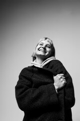 A woman with short hair smiles brightly while wrapped in a cozy coat, enjoying a peaceful moment. Soft lighting highlights her joyful demeanor and the simplicity of the scene
