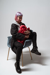 In a minimalist studio, a young person with pastel pink hair sits in a stylish chair, embracing a vibrant red blanket. The scene conveys a blend of comfort and emotion with a modern aesthetic