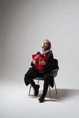 In a minimalistic setting, a person with vibrant hair sits in a stylish chair, holding a bright red gift. The atmosphere is filled with excitement and warmth, suggesting a special moment ahead