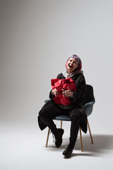 A woman with short pink hair sits on a stylish chair, joyfully holding a red gift close to her chest. Her laughter fills the minimalist space, creating a warm and cheerful atmosphere