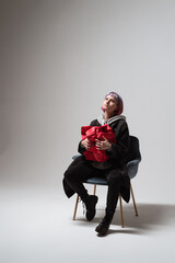 In a minimalist studio, a young person sits in a stylish chair, cradling a vibrant red object. Their expression reflects a deep contemplation, illuminated by gentle light
