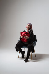 A woman wearing a casual outfit sits comfortably in a chair, cradling a vibrant red gift. Her expression radiates happiness as she enjoys this delightful moment of anticipation