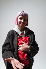 In a minimalistic studio, a smiling person with colorful hair holds a wrapped gift close to their chest. Their open hand suggests a welcoming gesture, enhancing the cheerful atmosphere