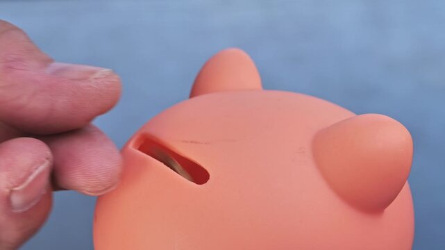 A hand carefully inserts a euro coin into the narrow slot of an orange, pig-shaped savings bank.