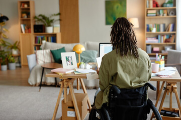 Young adult woman with disability sitting in wheelchair working at desk with computer in modern home office, surrounded by documents and personal items, facing away from camera
