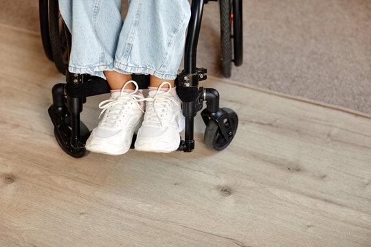 Woman sitting in wheelchair with disability, wearing sneakers and jeans, feet resting on footrests, lower legs and wheelchair frame visible on wooden floor