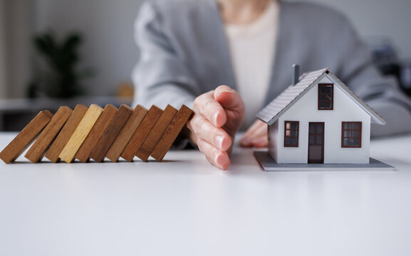 A woman's hand stops a wooden block from falling onto a model house. Home insurance and safety ideas and protecting your home from falling over wooden blocks.

 - Powered by Adobe