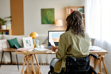Young adult woman with disability sitting in wheelchair, working at desk, using laptop and holding documents in modern home office with natural light coming through window