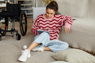 Young adult Caucasian woman with disability sitting on floor using digital tablet, wheelchair...
