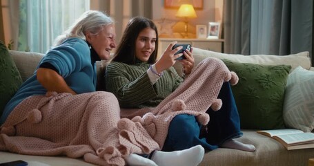 Grandmother and granddaughter laughing together while using funny mobile apps on a smartphone at home, enjoying family bonding and playful technology moments on the living-room sofa. - Powered by Adobe