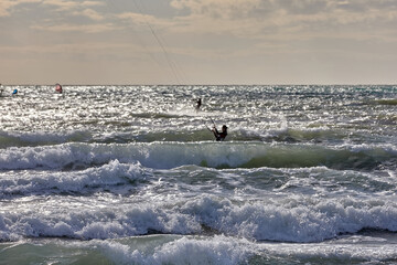 Kitesurfer glides through strong waves under an overcast sky, surrounded by the shimmer of sunlight on the restless sea. motion, energy