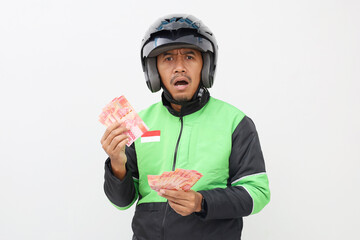 Shocked Asian online taxi driver guy holding Indonesian banknotes, white studio background.