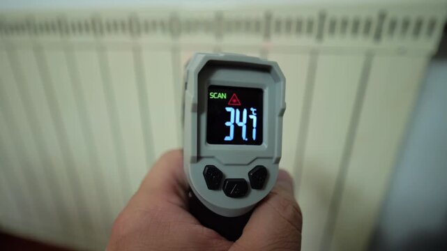 A hand holding an infrared thermometer aimed at a white radiator, demonstrating the process of checking thermal efficiency for central heating