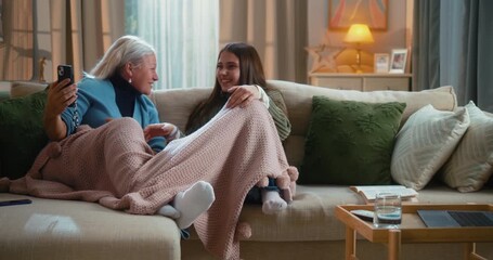 Grandmother and granddaughter laughing together while using funny mobile apps on a smartphone at home, enjoying family bonding and playful technology moments on the living-room sofa. - Powered by Adobe