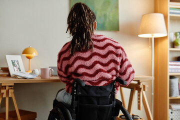 Young adult woman sitting in wheelchair working at desk with laptop in home office setting, long dreadlocks visible, back facing camera, modern workspace environment