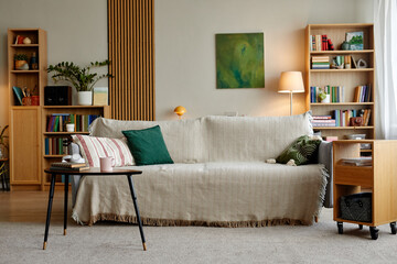 Modern living room interior featuring beige sofa with decorative pillows, wooden bookshelves filled with books and plants, coffee table with headphones and mug, cozy home setting