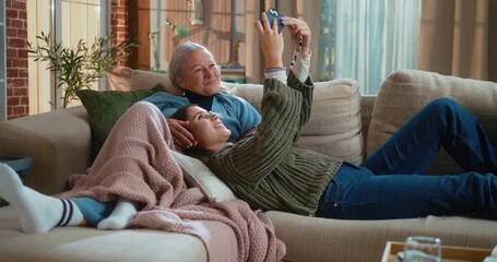 Teen granddaughter lying in her grandmother’s lap and taking a selfie together on the living room sofa at home, capturing a warm family bonding moment for social media. - Powered by Adobe