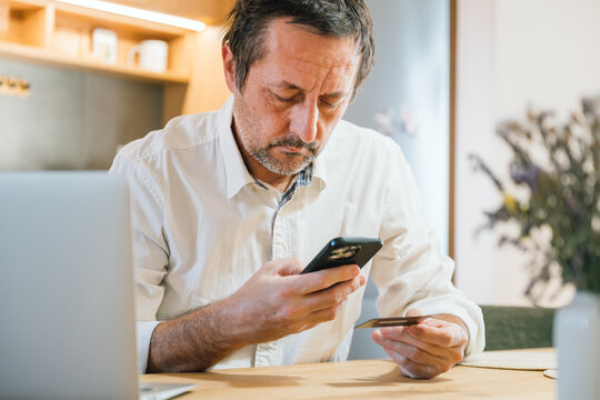 Unshaved businessman sitting at desk in home office, holding credit card and smartphone, online payment concept.