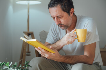 Serious mature man reading a yellow hardcover book in a cozy living room in the morning, holding a hot drink and focusing deeply.