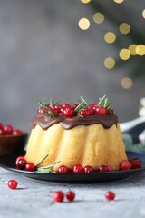 Delicious Christmas cake with chocolate glaze, cranberries and rosemary on grey textured table against blurred lights, closeup. Bokeh effect