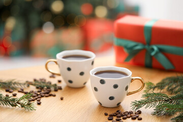Aromatic coffee in cups, fir tree branches, beans and gift box on wooden table against blurred lights, closeup. Christmas greeting card