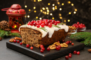 Tasty Christmas cake with icing, cranberries, nuts and festive decor on black table against blurred lights, closeup. Bokeh effect