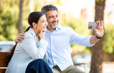 Happy mature couple taking a selfie together on a park bench on a sunny day
