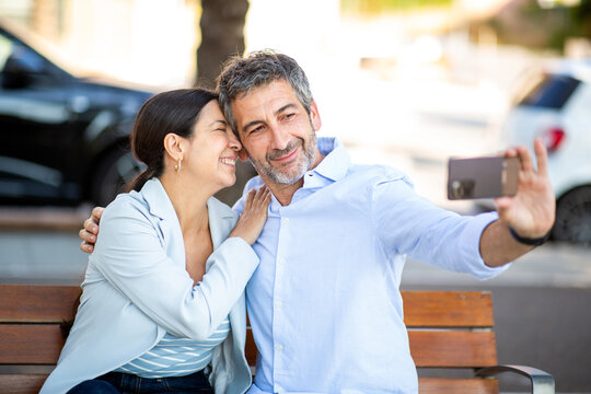 Affectionate couple smiling and cuddling while taking a selfie on bench - Powered by Adobe