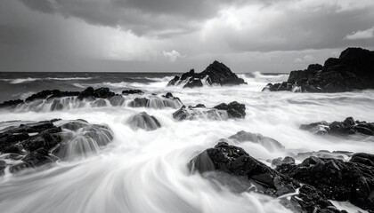 A dramatic black and white long exposure photograph captures the raw power of the ocean as waves crash against jagged rocks under a moody, cloudy sky.