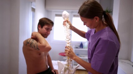 Female doctor showing a tattooed male patient with neck and shoulder pain a model of the human spine, explaining the cause of his discomfort during a consultation in a modern medical clinic