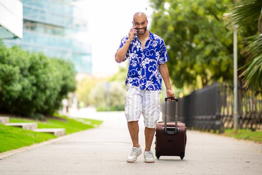 Smiling man in floral shirt on phone call while walking with suitcase outdoors